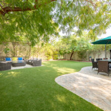 Real estate photography of a private back garden with green lawn, mature trees, patio furniture, and parasol