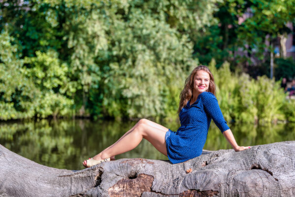 Candid portrait of woman in blue dress seated on a log by a Vondelpark pond, Amsterdam