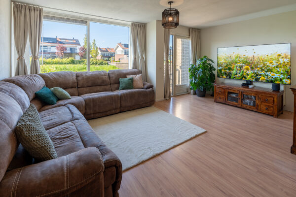 Real estate photography of a sunlit living room with sofa, plants, and full garden view through tall windows