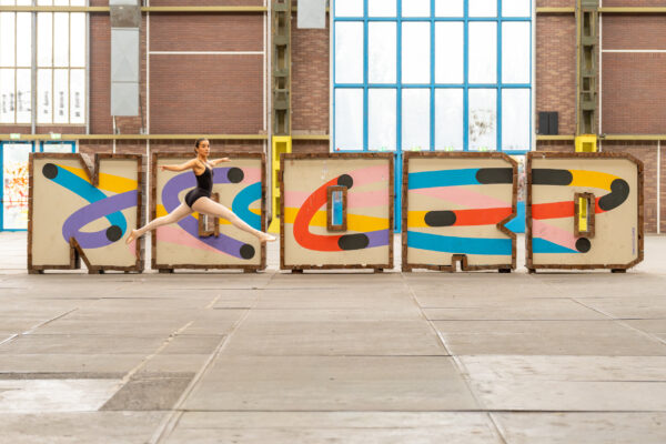 Ballet dancer mid-leap in front of the colourful "Noord" sign inside an NDSM warehouse, Amsterdam Noord