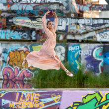 Ballet dancer in pink dress and pointe shoes mid-leap against NDSM graffiti wall, Amsterdam Noord