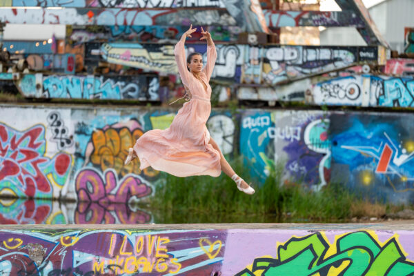 Lifestyle photographer Amsterdam — ballet dancer in pink dress and pointe shoes mid-leap against NDSM graffiti, Amsterdam Noord