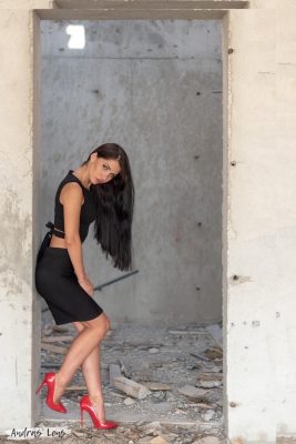 Fashion model standing against the wall at a construction site in Dubai, wearing red heels, black skirt and top.