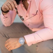 Photograph of the upper body only of a fashion model in Dubai wearing a pink shirt, watch while holding a pair of sunglasses.